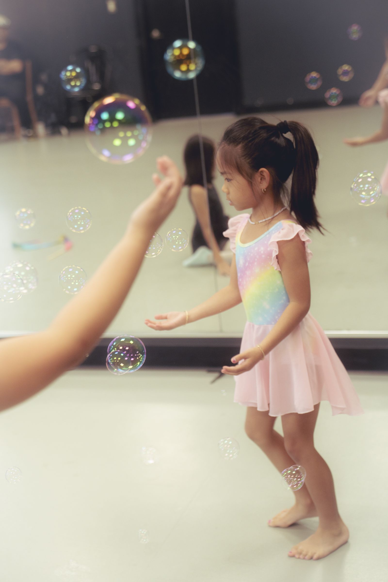 A young dancer reaches out to catch floating soap bubbles in front of the studio mirror.