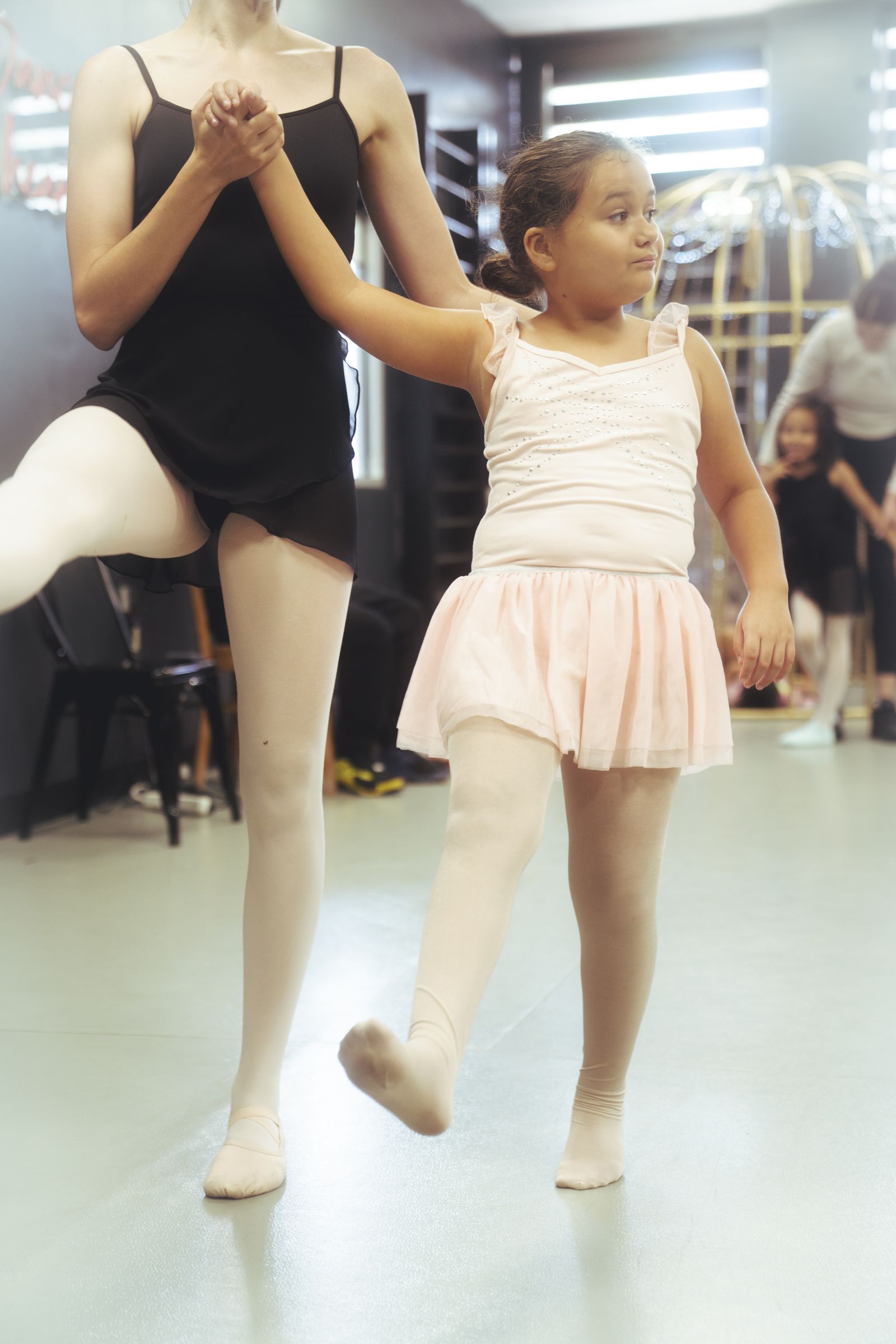An instructor holds a young dancer's raised hand for balance during a ballet step.