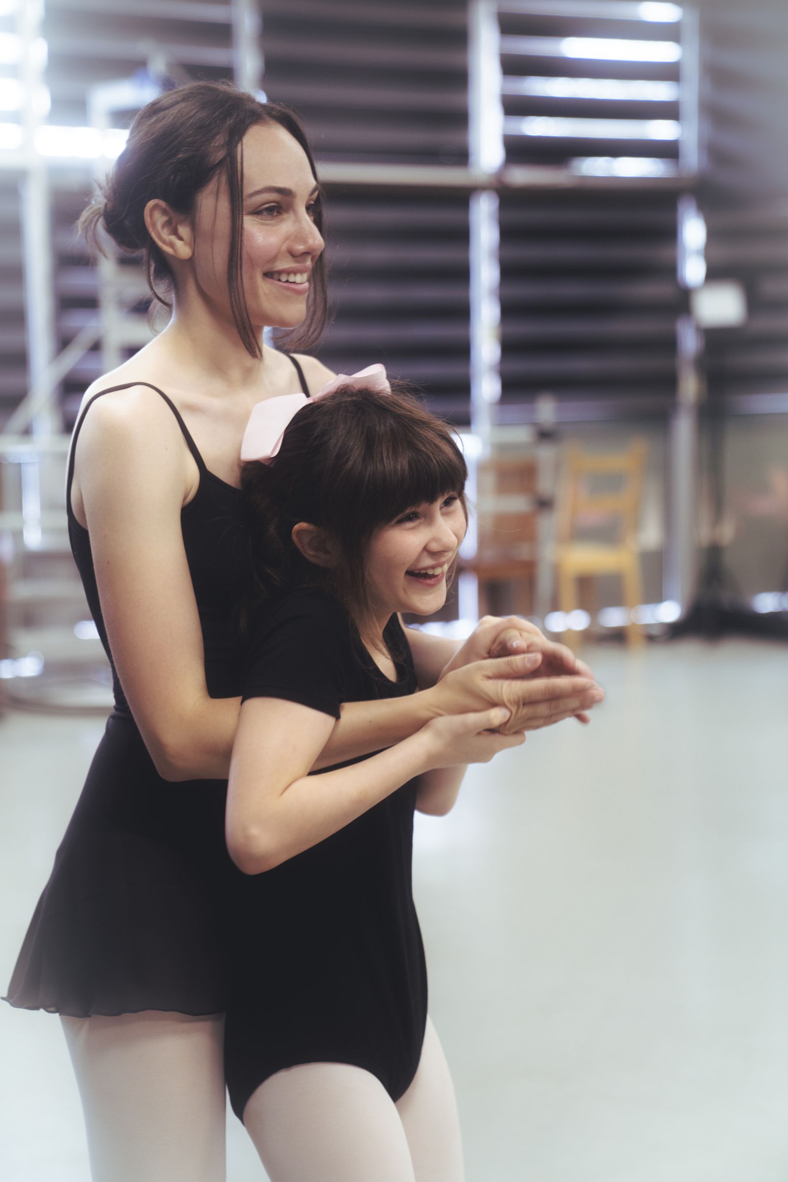 An instructor and a young dancer laugh together during class, their hands clasped.