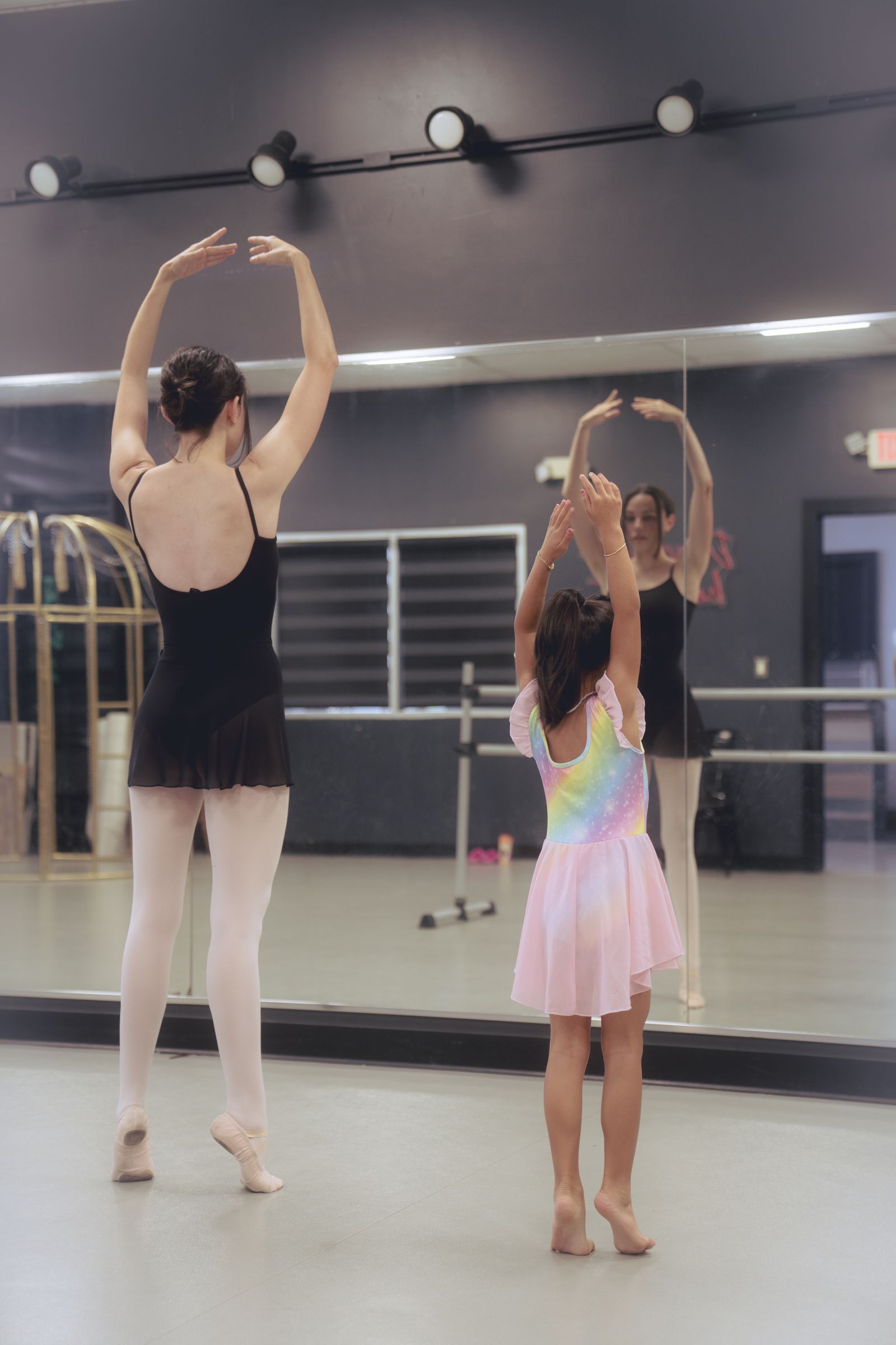 A young dancer raises both arms overhead in front of the studio mirror, where the instructor's reflection echoes the same pose.